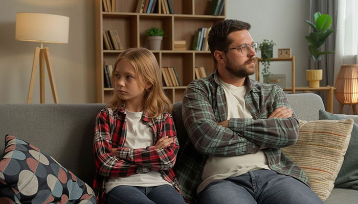 Father and teenage daughter sitting apart on couch with arms crossed, showing tension over paper towel swimming idea on period.