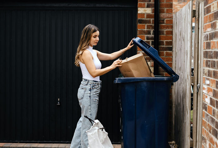 Woman placing a cardboard box in a trash bin outside a brick house, demonstrating moving advice with boxes and packing.