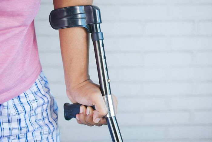 Person holding a metal crutch with an adjustable height mechanism against a white brick wall background.