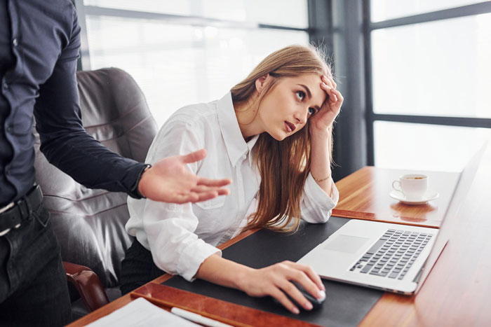 Frustrated woman at desk with laptop looking stressed while coworker gestures, illustrating work life made insufferable.