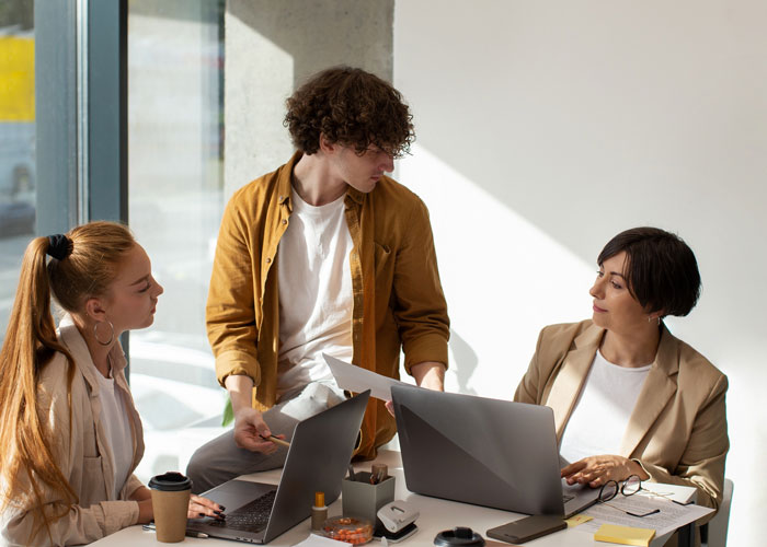 Three coworkers in a meeting, one appearing to push beliefs while others look uncomfortable at a modern office table.