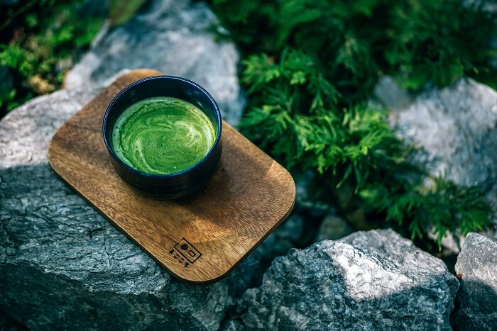 A bowl of frothy green matcha tea on a wooden tray placed on rocks with greenery in the background for unpopular opinions poll.