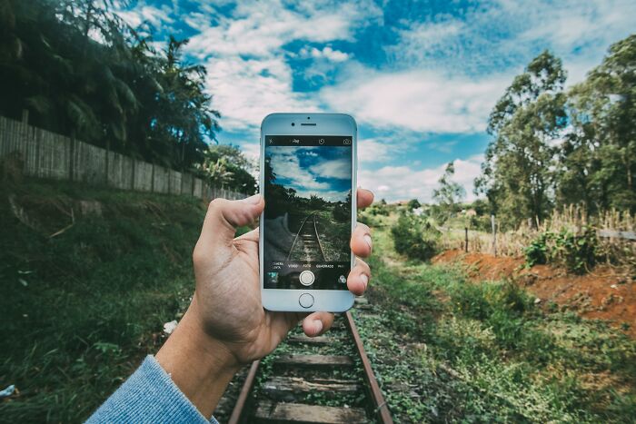 Hand holding smartphone capturing railroad track under blue sky, illustrating unpopular opinions poll concept outdoors.