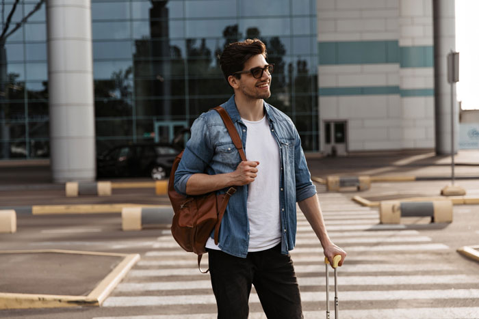 Young man with sunglasses and backpack standing outdoors with luggage, symbolizing surprise family visit during surgery planning.