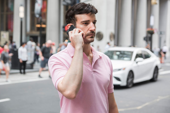 Man in pink shirt on phone outdoors in city, appearing concerned about surprise family visit during surgery plans.