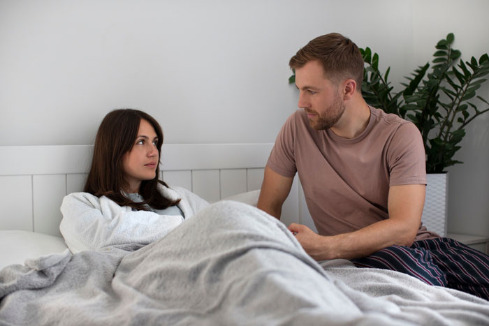 Couple in bedroom with woman resting under blanket after surgery while boyfriend sits beside her showing concern.