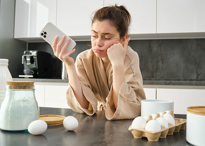Bride with allergy looking disappointed while holding phone in kitchen, surrounded by baking ingredients, no cupcakes from wedding vendors.