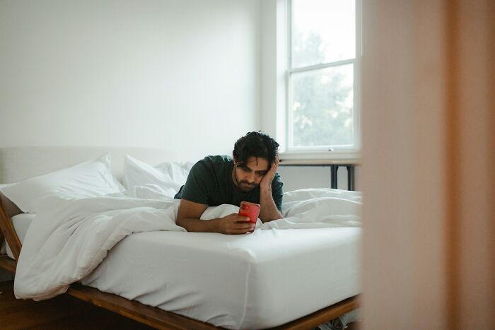 Man lying on bed looking at phone, reflecting on chaotic breakup stories shared by exes in a bright bedroom.