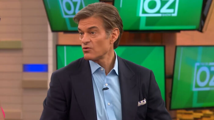 Man in a dark suit and blue shirt speaking on a health show set with multiple green screens displaying the word Oz.