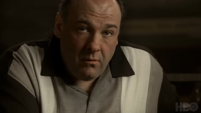 Man in a dimly lit room, wearing a black and white shirt, appearing thoughtful.