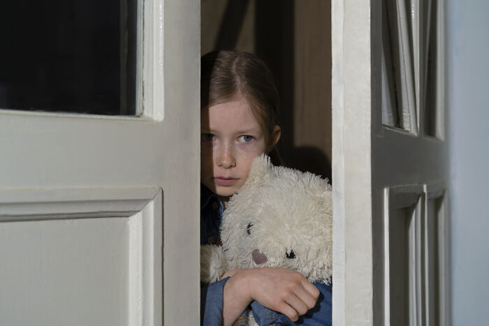 Young girl holding a teddy bear peeks anxiously from behind a partially open door in a dimly lit room, creepy experience theme.