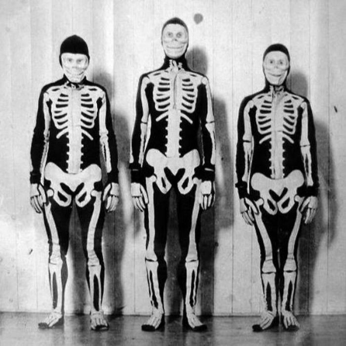 Three people wearing vintage skeleton Halloween costumes standing against a plain wall, showcasing classic terrifying designs.
