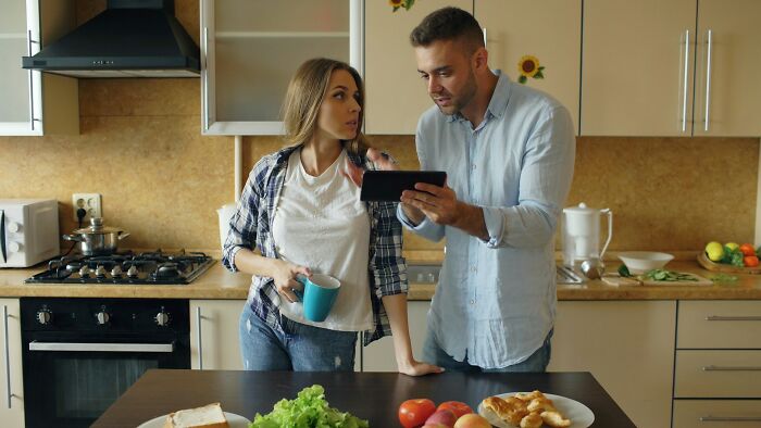 Man showing phone to woman in kitchen, discussing possible cheating signs in women from a broken-hearted man's perspective.