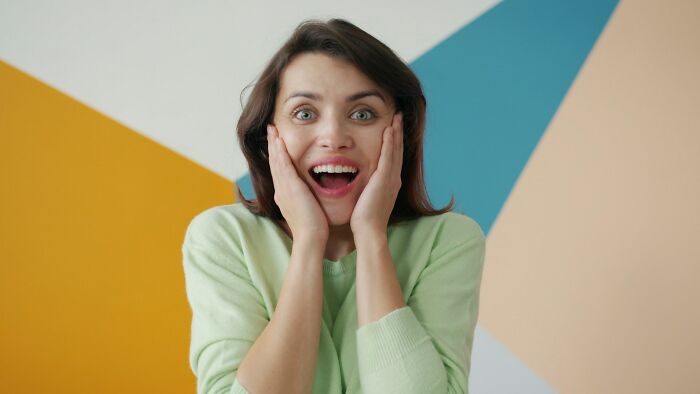 A woman in a green sweater with hands on cheeks, expressing surprise and delight, illustrating karma striking moments.