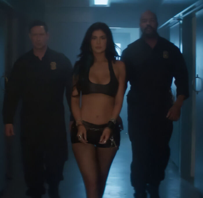 Kylie Jenner walking handcuffed between two officers in a dimly lit corridor in a fake arrest video amid ICE raids controversy.