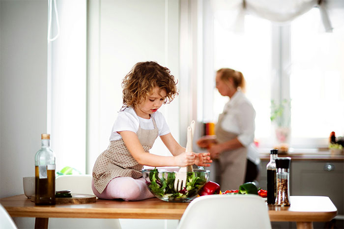 Young child wearing apron mixing salad on table while adult in background cooks, illustrating cooking smarter kitchen hacks.