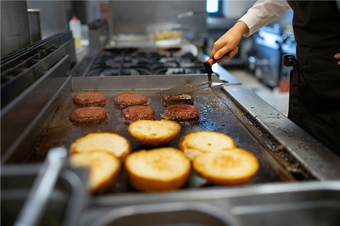 Person cooking burger patties and toasting buns on a large grill, demonstrating smart kitchen cooking cheats.