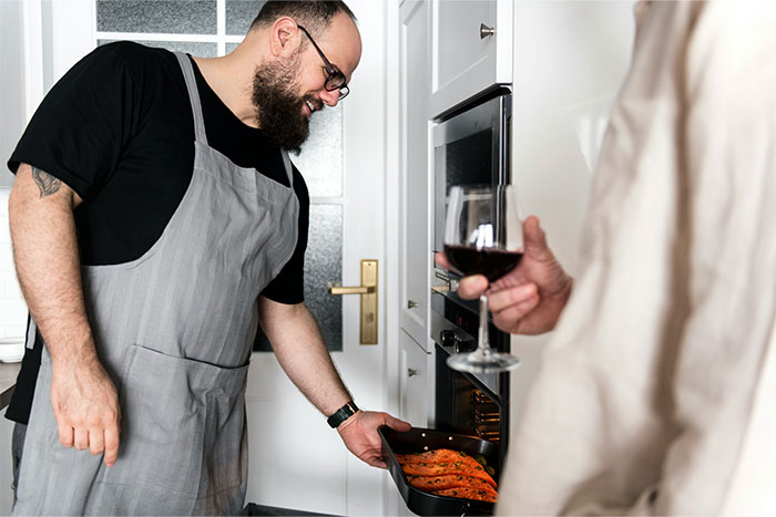 Man in a grey apron cooking salmon in an oven while another person holds a glass of red wine in a modern kitchen.