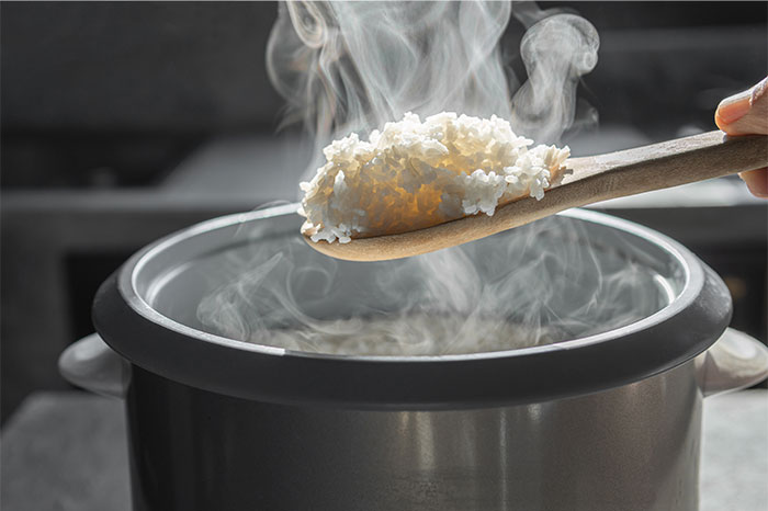Steaming cooked rice being scooped with a wooden spoon from an electric rice cooker in a home kitchen.