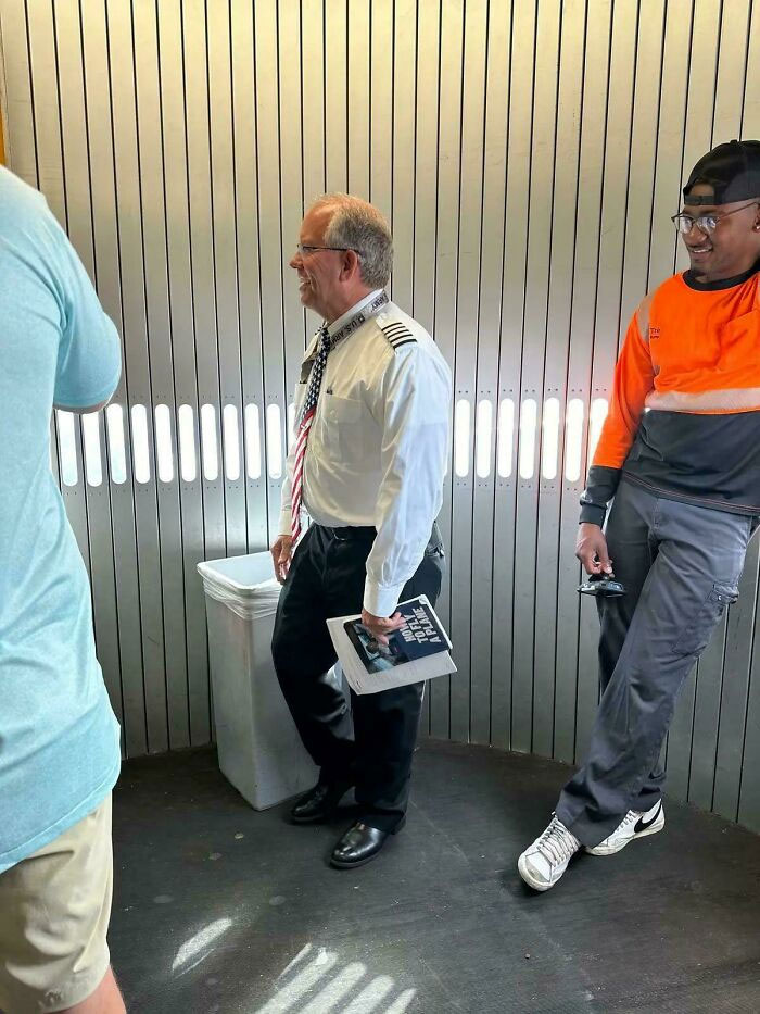 Pilot in uniform holding papers talking to casually dressed man in orange safety shirt inside metal-paneled room.