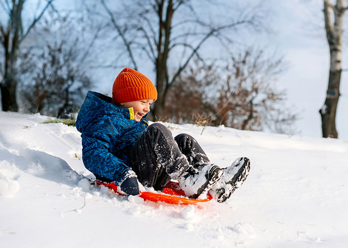Niño feliz con gorro naranja y abrigo azul deslizándose en trineo sobre nieve, imagen relacionada con hallazgos autopsia médicos.