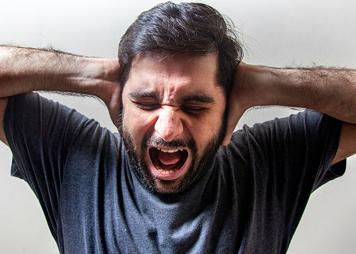 Man with beard and dark hair holding his head and screaming, expressing distress related to medical examinations uncovering truths.