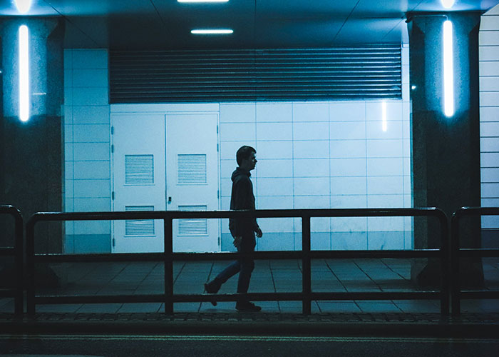 Silhouetted man walking alone at night near railing and illuminated wall, evoking medical examinations uncovering disturbing truths.