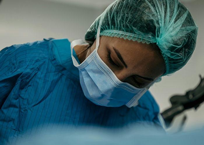 Medical examiner wearing surgical mask and cap performing a detailed medical examination in a clinical setting.