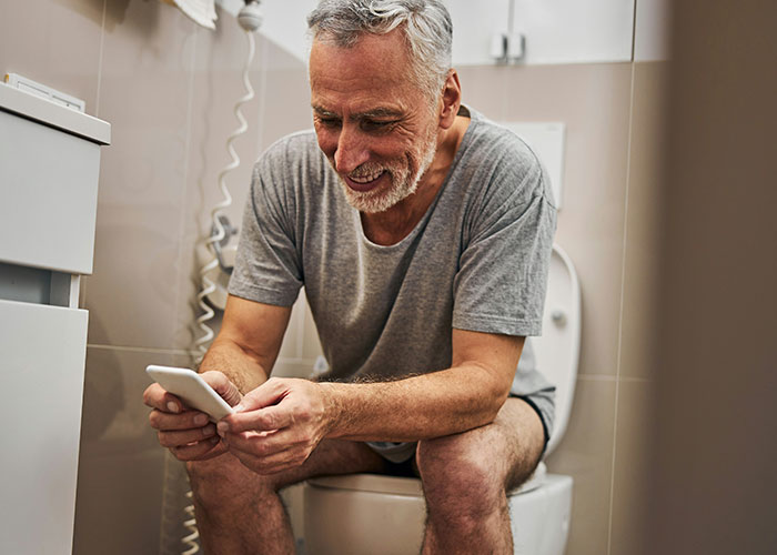 Hombre mayor sentado en el baño usando teléfono móvil, relacionado con hallazgos sorprendentes de médicos forenses.