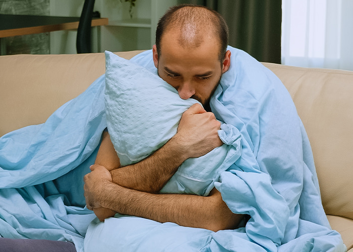 Man wrapped in blue blanket hugging a pillow, showing stress and fatigue in a dad in maternity wards moment.