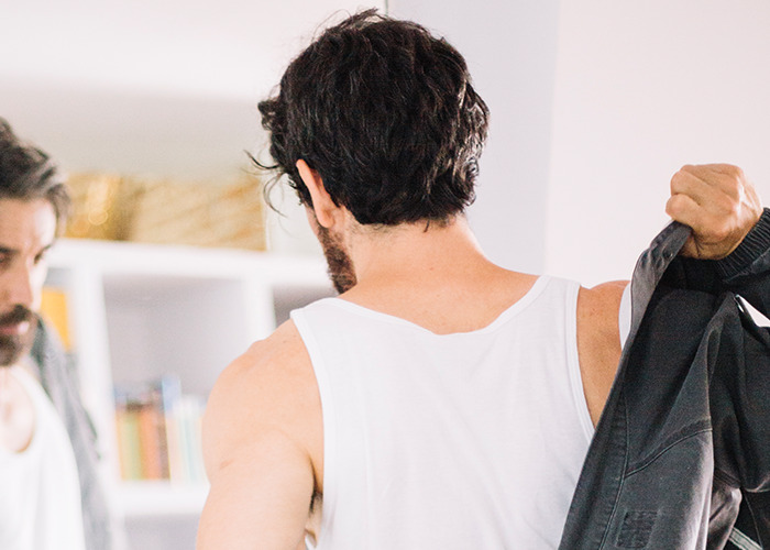 Man in a white tank top putting on a dark jacket, reflecting in a mirror, capturing dad in maternity wards moment.