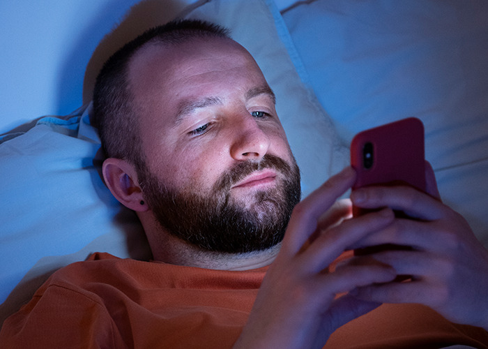Bearded man lying in bed at night, focused on his phone, illustrating dad in maternity wards stories humor and frustration.