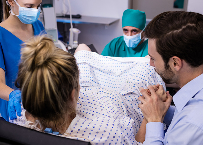 Father anxiously supporting partner during childbirth in maternity ward with medical team assisting delivery.