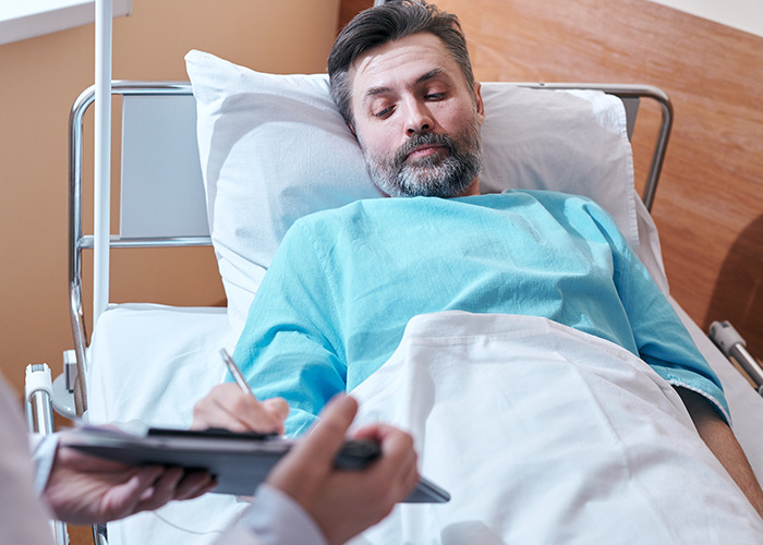 Man in hospital bed writing notes while interacting with medical staff, illustrating dad in maternity wards experience.