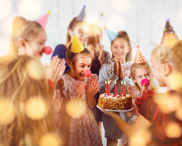 Group of children wearing party hats and clown noses celebrating birthday with cake and candles indoors