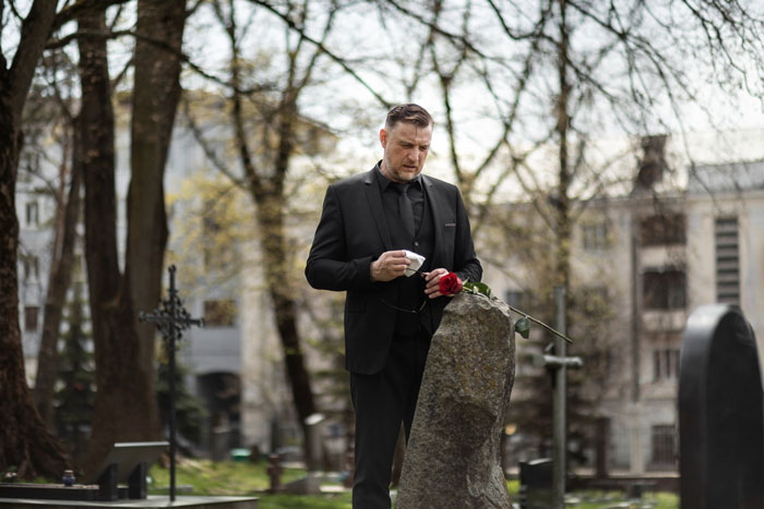 Man in black suit holding a red rose at a gravestone, reflecting on dying wife&rsquo;s wishes about dating after loss.