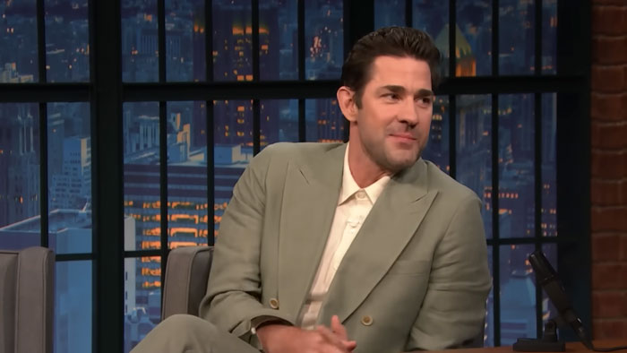John Krasinski in a gray suit smiling during a nighttime talk show interview in a studio setting.