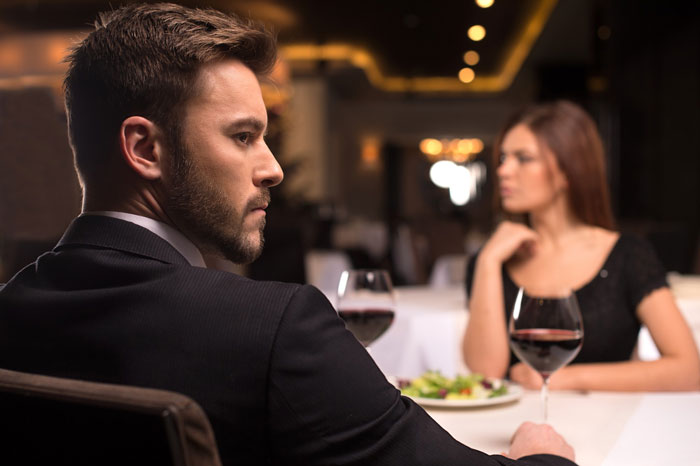 Man in suit looking upset while woman sits across the table at a restaurant, illustrating mad hubs and free pass with John Krasinski.