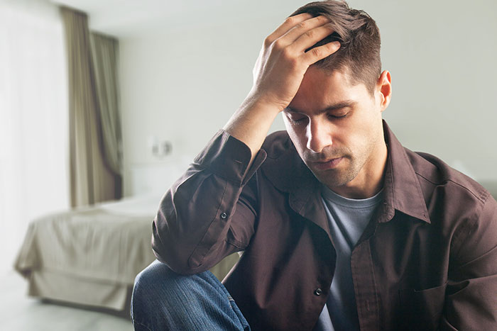 Man sitting on the floor in a bedroom, looking distressed and holding his head, reflecting anger and regret.