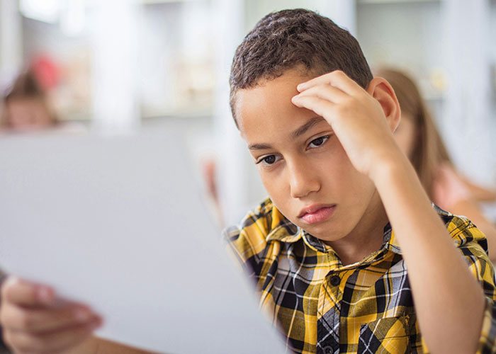 Boy in a yellow plaid shirt showing a thoughtful expression while holding a paper, capturing moment of instant karma manifestations.