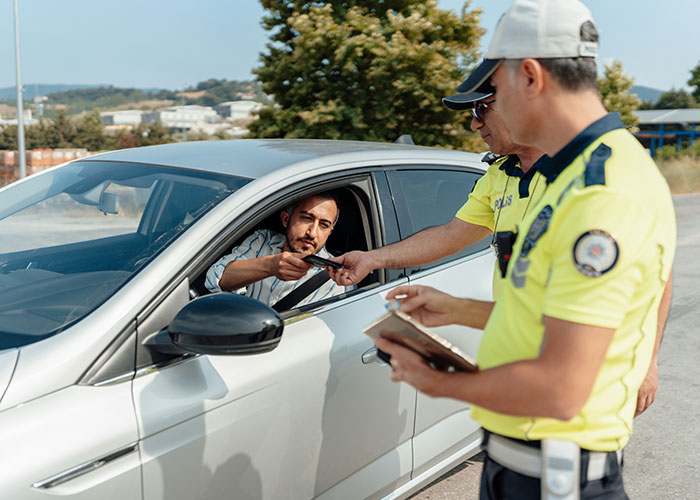Man in car handing documents to police officers during instant karma traffic stop on a sunny day outdoors.