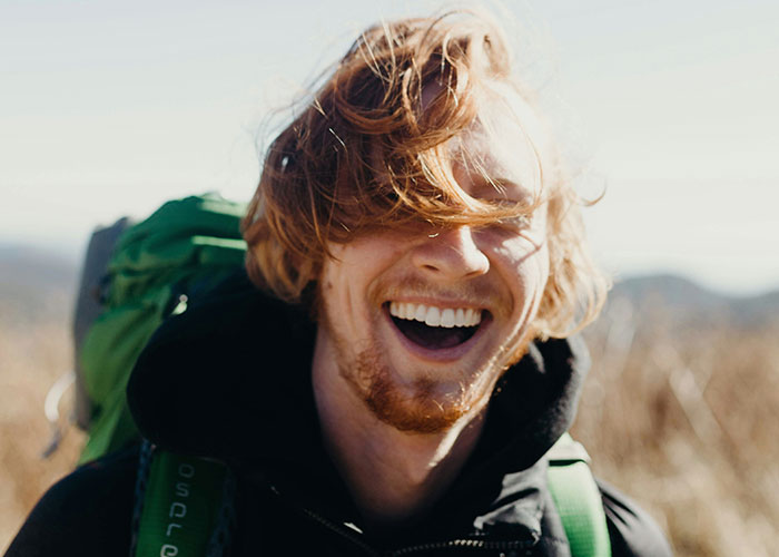 Young man with red hair and a backpack laughing outdoors, capturing a moment of glorious instant karma manifestation.