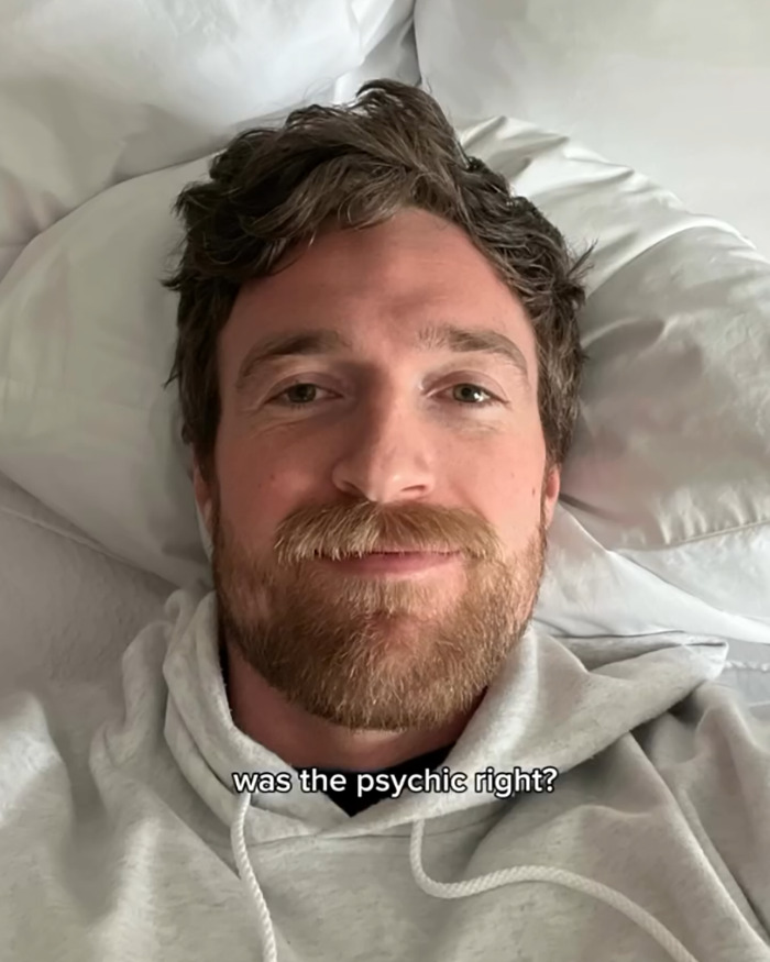 A bearded man lying on a bed wearing a gray hoodie with text about a psychic drawing changing love life.