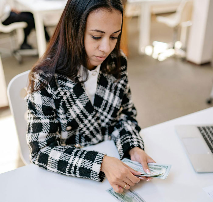 Upset woman counting money at desk, reflecting shock after bride-to-be asks her to pay for bridesmaid dress.