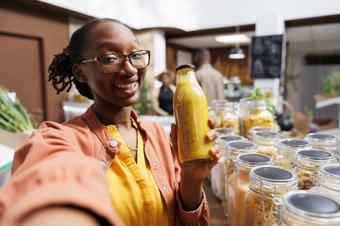 Smiling woman holding a bottle of healthy dressing in a kitchen with jars of ingredients, promoting healthy habits.