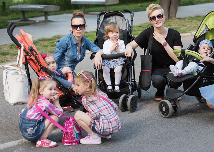 Two women with two kids each in strollers, struggling for support after surgery while caring for their children outdoors.