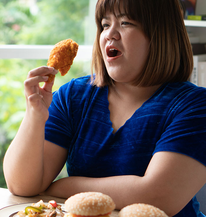 Woman holding fried chicken, sitting at table with burgers, illustrating bill splitting and fat-shaming friends discussion.