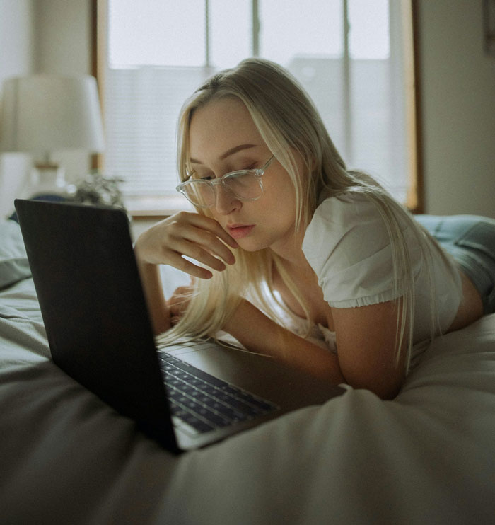 Young woman with glasses lying on bed looking at laptop screen, appearing thoughtful and concerned about relationship news.