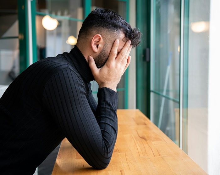 Man covering face with hands sitting alone by window showing distress in a cafe, reflecting emotional turmoil in relationship.