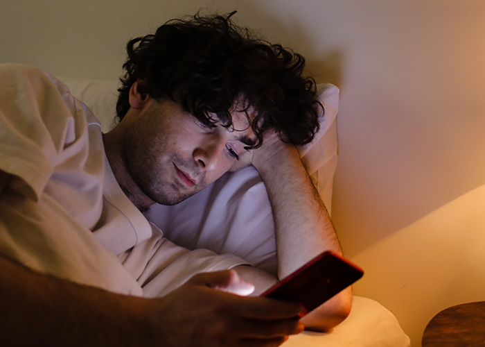 Young man lying in bed using smartphone at night, reacting to story about inappropriate Halloween costume conflict.
